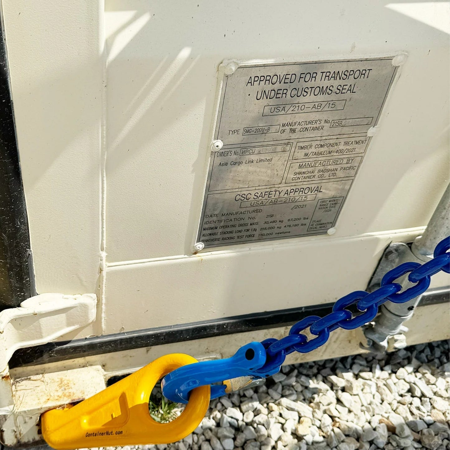 Close-up of a yellow locking mechanism (Right/Left hook) securing a corner of a white container with the Chain Sling in blue attached. The container is resting on a concrete block, and there is a metal identification plate on its side.