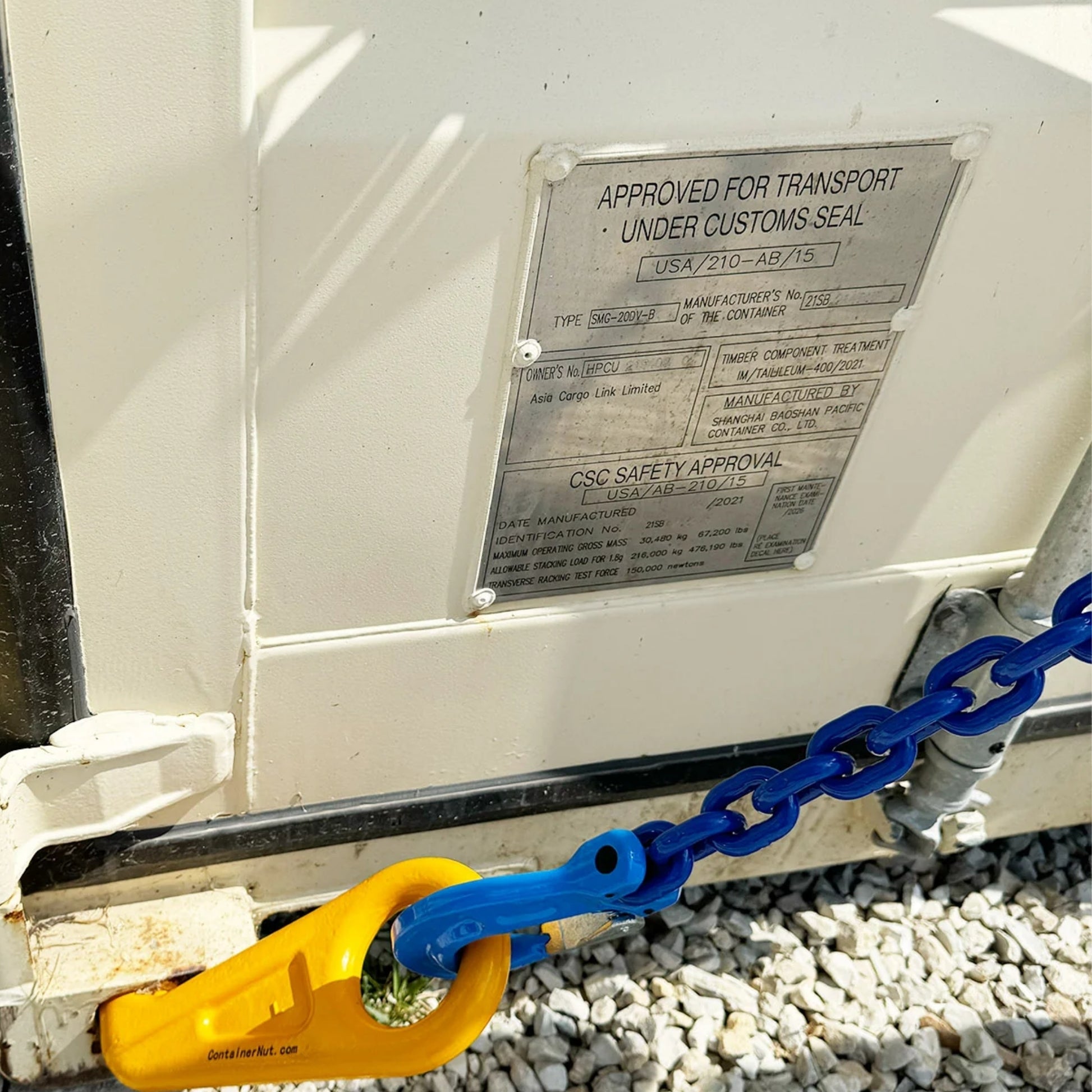 Close-up of a yellow locking mechanism (Right/Left hook) securing a corner of a white container with the Chain Sling in blue attached. The container is resting on a concrete block, and there is a metal identification plate on its side.