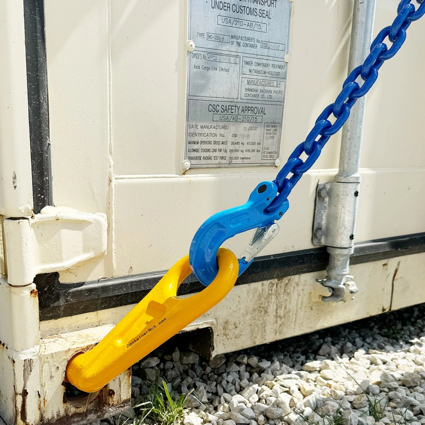 Close-up of a yellow locking mechanism (Right/Left hook) securing a corner of a white container with the Chain Sling in blue attached. The container is resting on a concrete block, and there is a metal identification plate on its side.