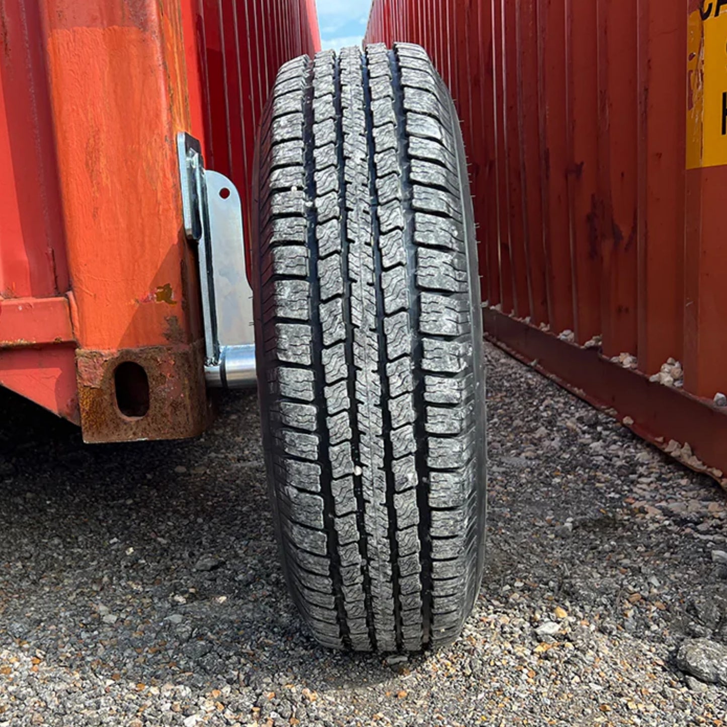 A close-up of a tire using EZY Wheels by Container Nut situated between two red shipping containers on gravel ground.