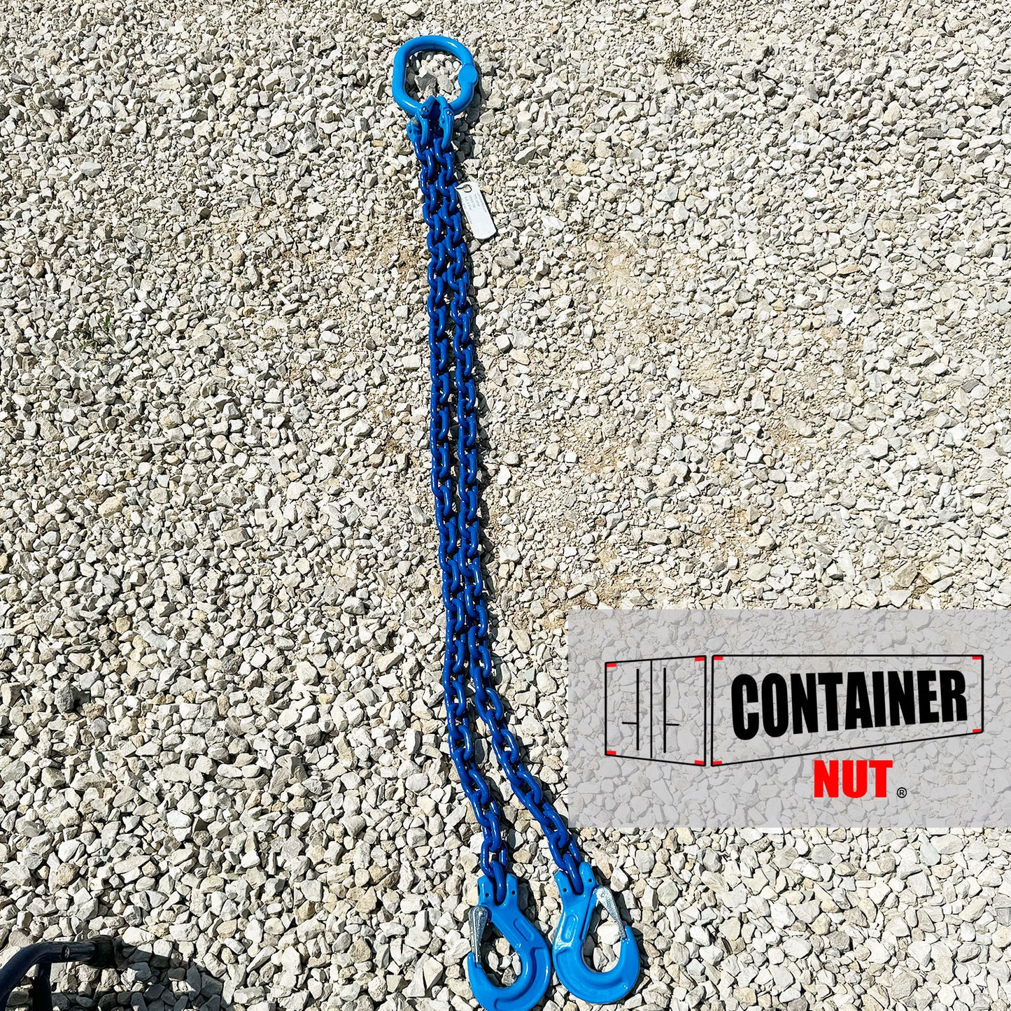 A Chain Sling from Container Nut, featuring a blue chain with two hooks and a circular ring, is laid out on a gravel surface. A partially visible label is attached to the chain.