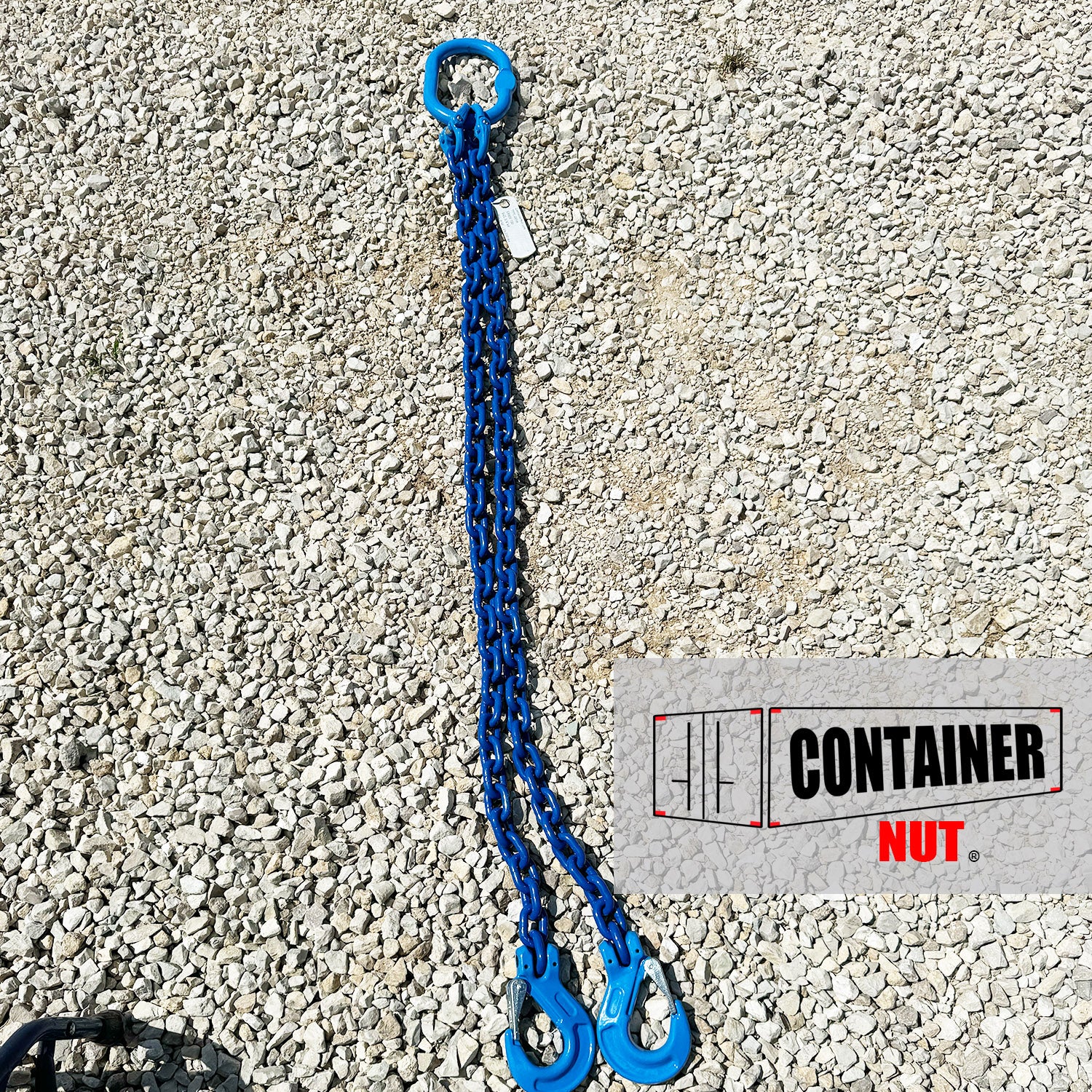 A Chain Sling from Container Nut, featuring a blue chain with two hooks and a circular ring, is laid out on a gravel surface. A partially visible label is attached to the chain.