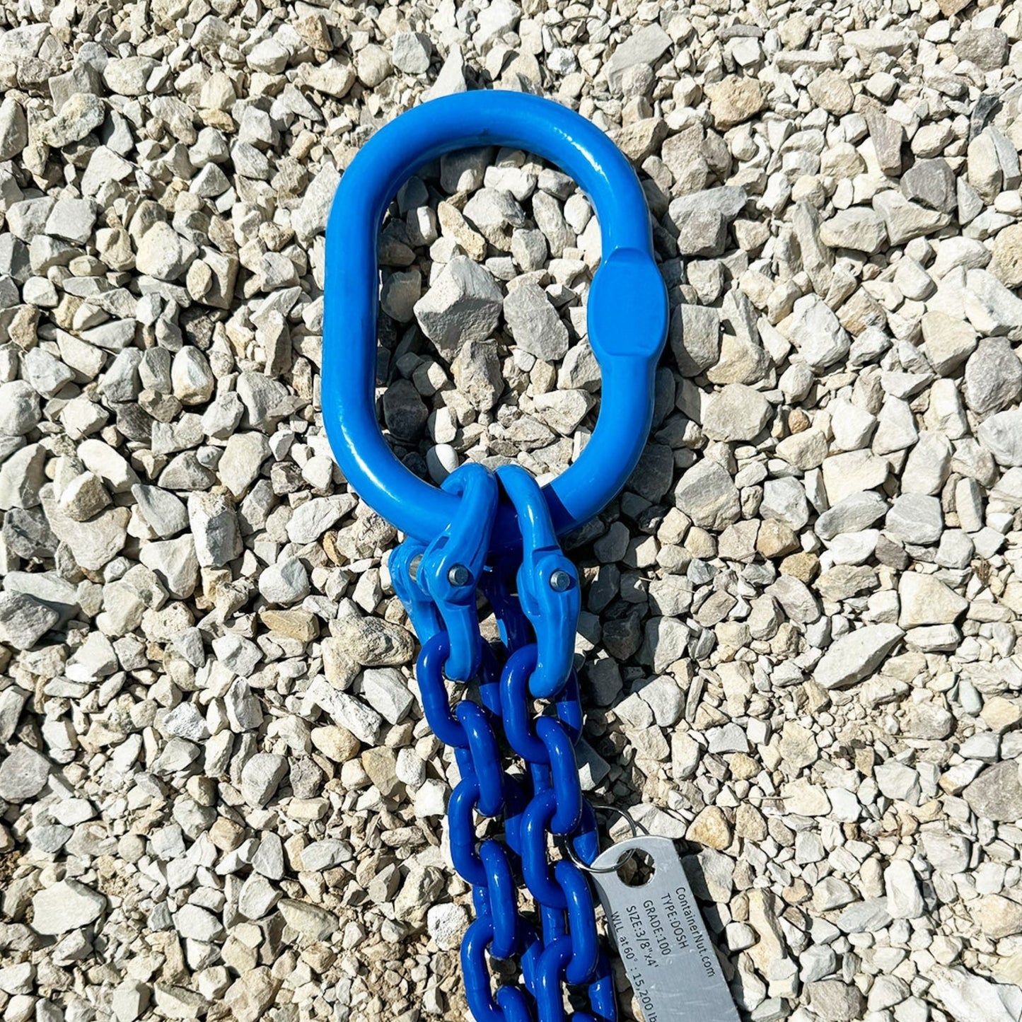 Bright blue metal Chain Sling with an oval-link connector is laid out on gravel. Attached to the chains is a metal tag.