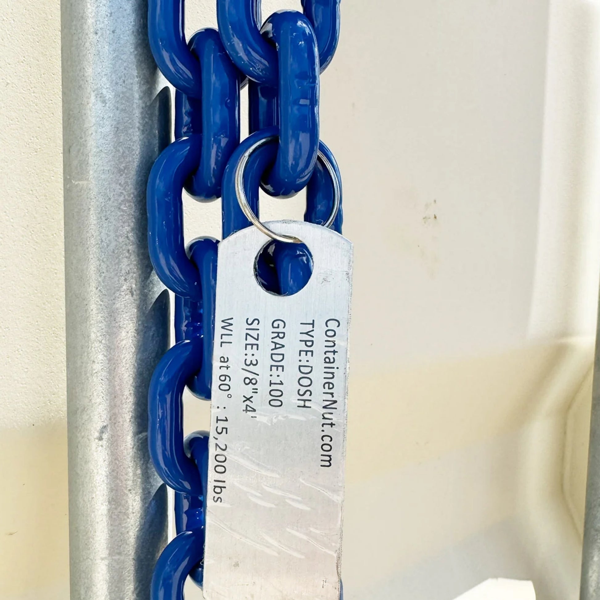 Close-up of a metal identification tag attached to a blue chain. The tag reads: "Containernut.com, TYPE: DOSH, GRADE: 100, SIZE: 3/8” x 4, WLL at 60° - 15,200 lbs." This is part of the Chain Sling + Right/Left Hooks Bundle from Container Nut.