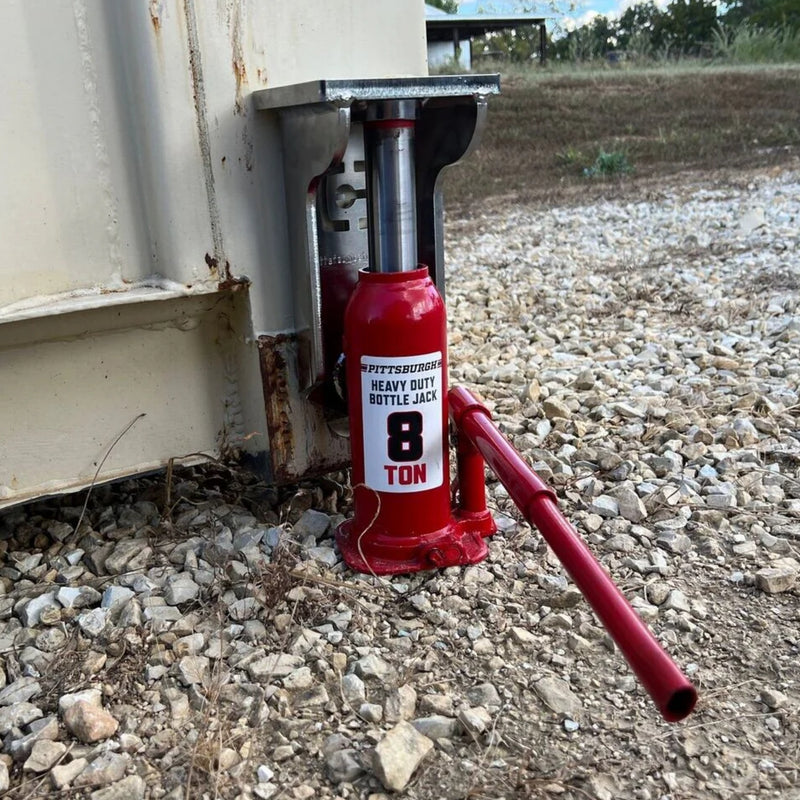 Load image into Gallery viewer, An EZY Lift, with a silver finish, is being used with a bottle jack to lift a container. It is placed on gravel next to the base of a beige outdoor structure. The detached handle lies on the ground beside the jack. The background features a grassy area and some trees.
